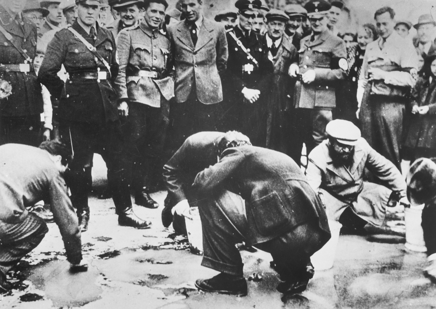 Jewish men kneeling on a Vienna pavement scrubbing it while a crowd of Austrians stands watching.