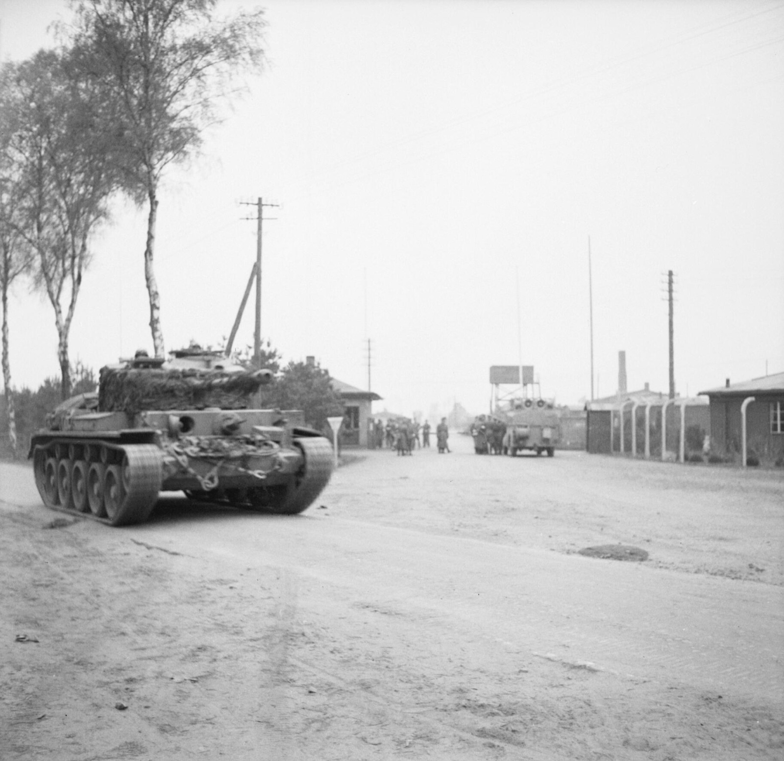 A tank moves along a track past a wooden camp gateway and fencing, with a group of figures standing at the roadside.