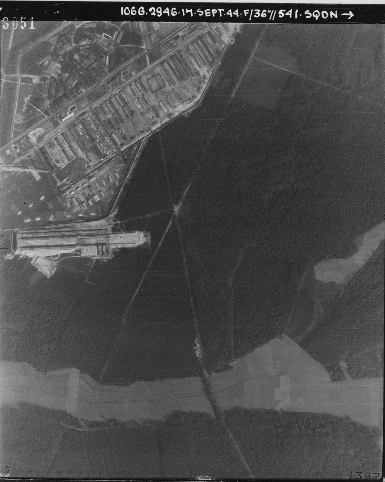 A black and white aerial photograph of a rectangular camp: parallel rows of long huts, straight service roads, open squares between the blocks, heath and forest around the perimeter.