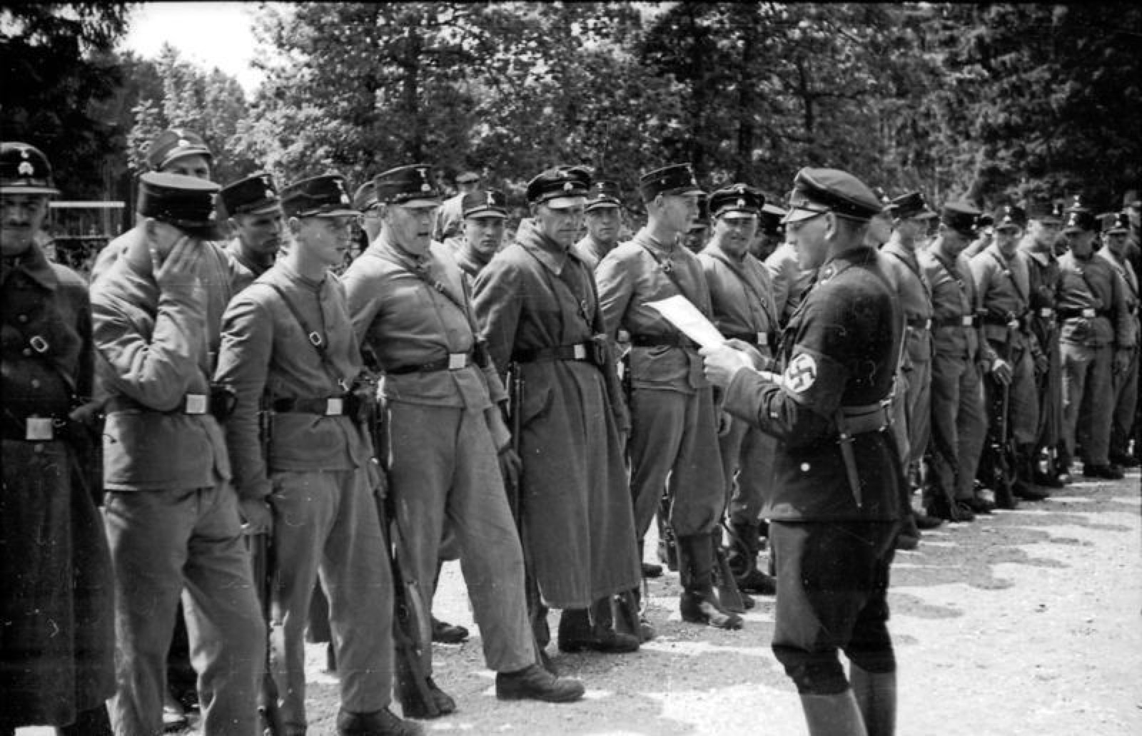 A long line of uniformed SS men at attention in ranks on open ground, photographed from one end so the row recedes into the distance, low camp buildings behind them.