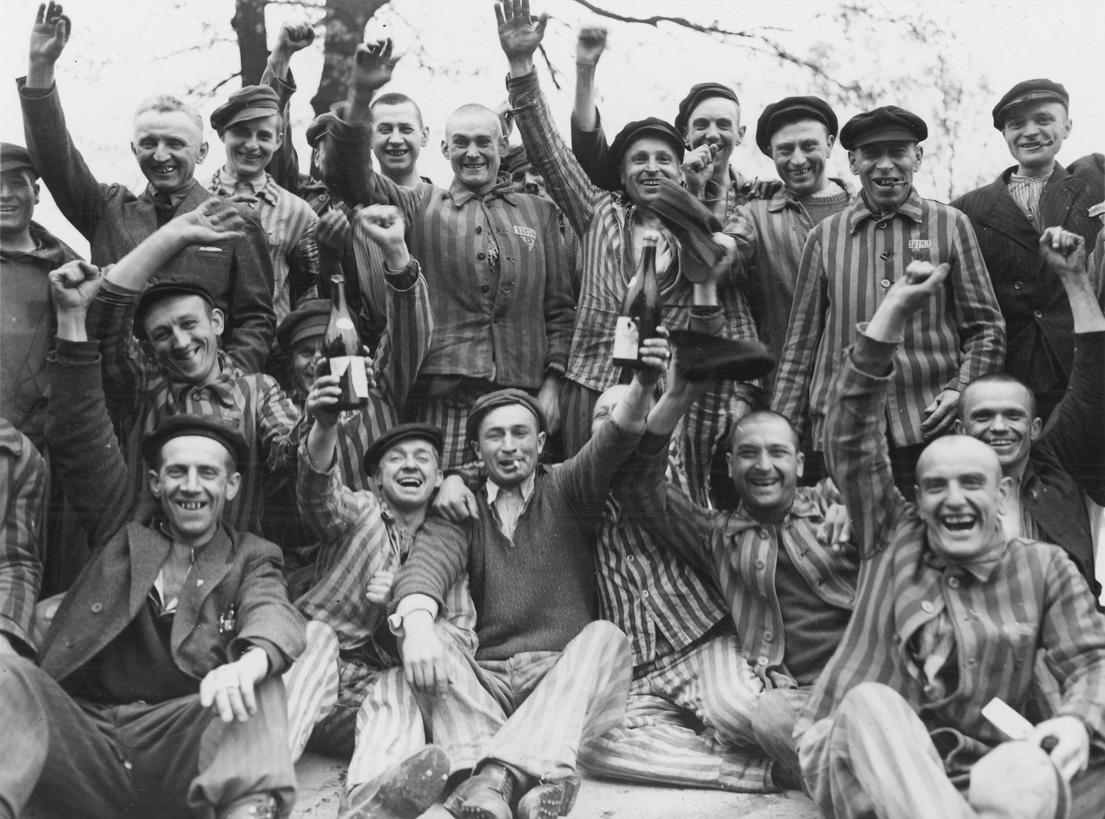 A crowd of thin men in prison stripes and mixed clothing pressed together indoors, arms raised, several holding bottles up toward the camera.