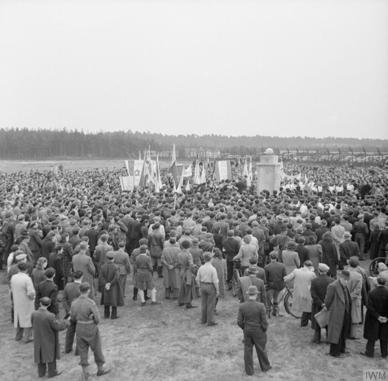 A crowd stands bareheaded around a low stone monument in an open field, several men holding the corners of a covering cloth.