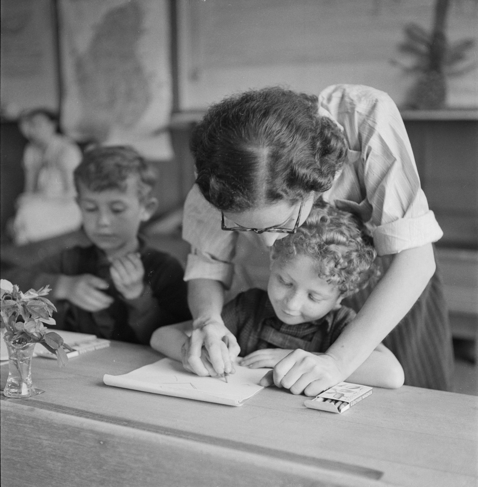 Children sit at wooden desks in a bare room writing in exercise books while a woman leans over one of them.