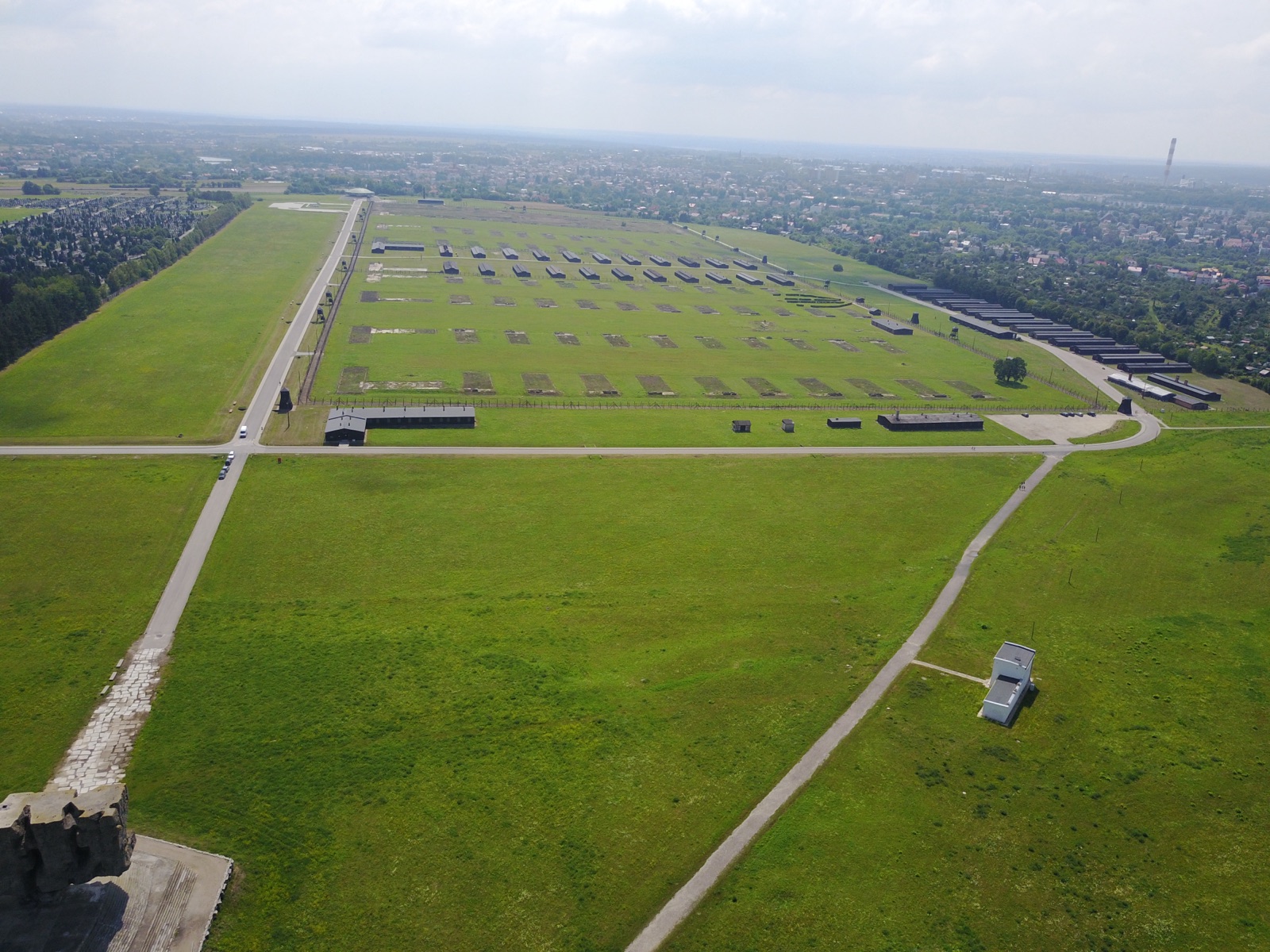 A color aerial photograph of long low wooden barracks in ordered rows, ringed by fencing, with a chimney and a domed structure at one edge.