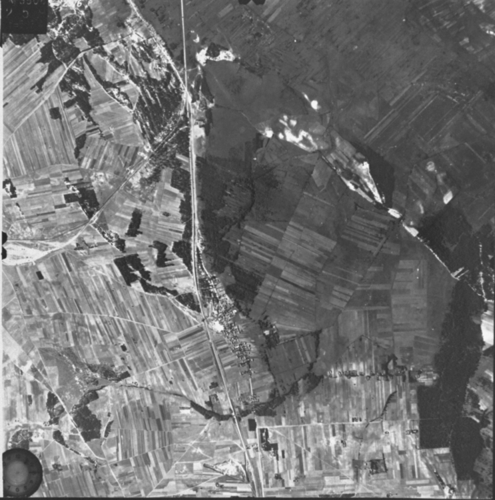 A grainy black and white air photograph of open country crossed by field boundaries and a rail line, with no camp structures visible.