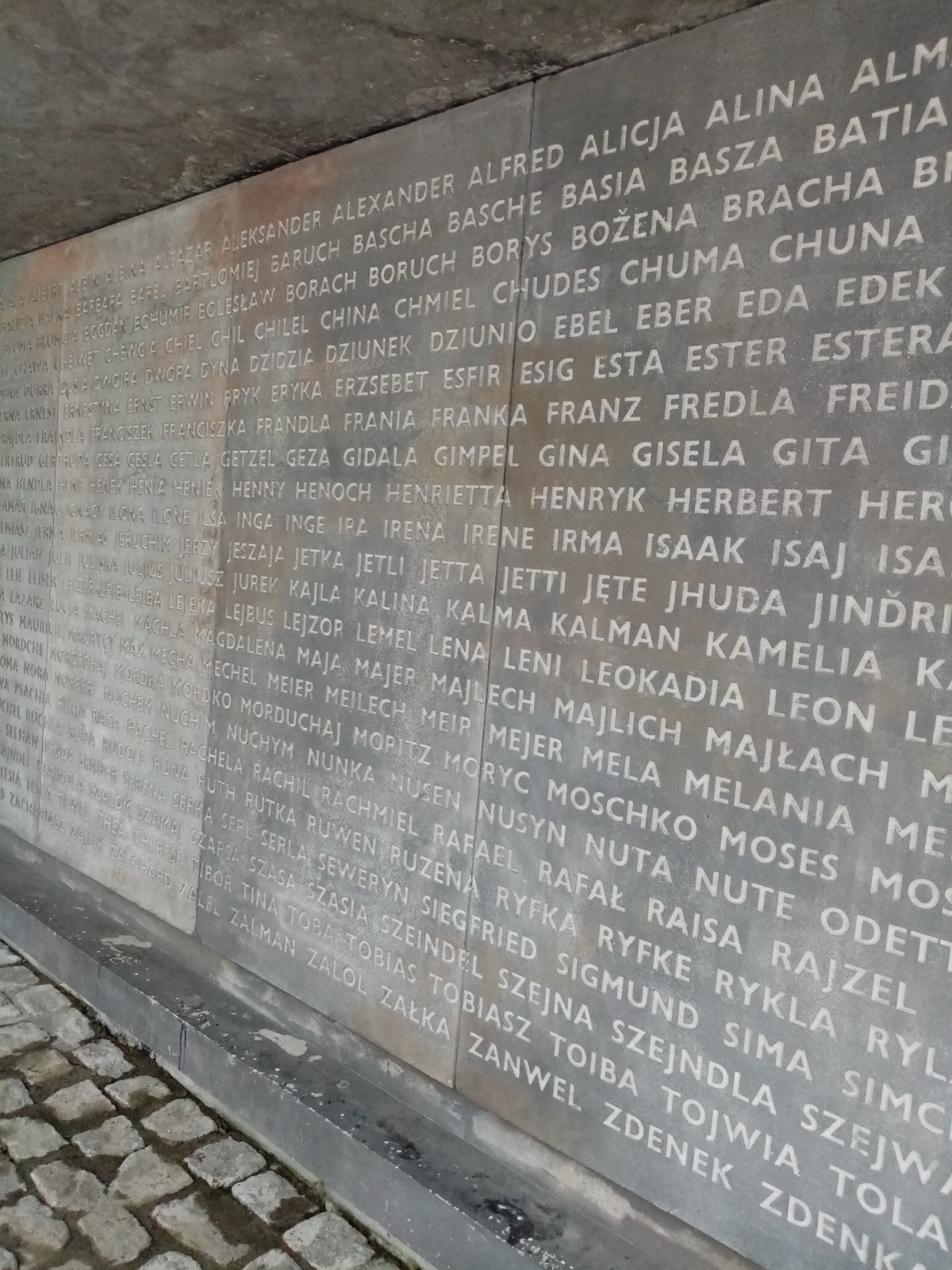 A tall dark stone wall cut with rows of Hebrew and Latin-script first names, lit from one side, filling the frame.