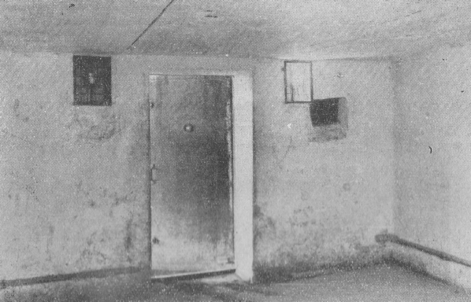 An empty concrete room with a heavy fitted door, a pipe entering high on one wall, and staining across the plaster.