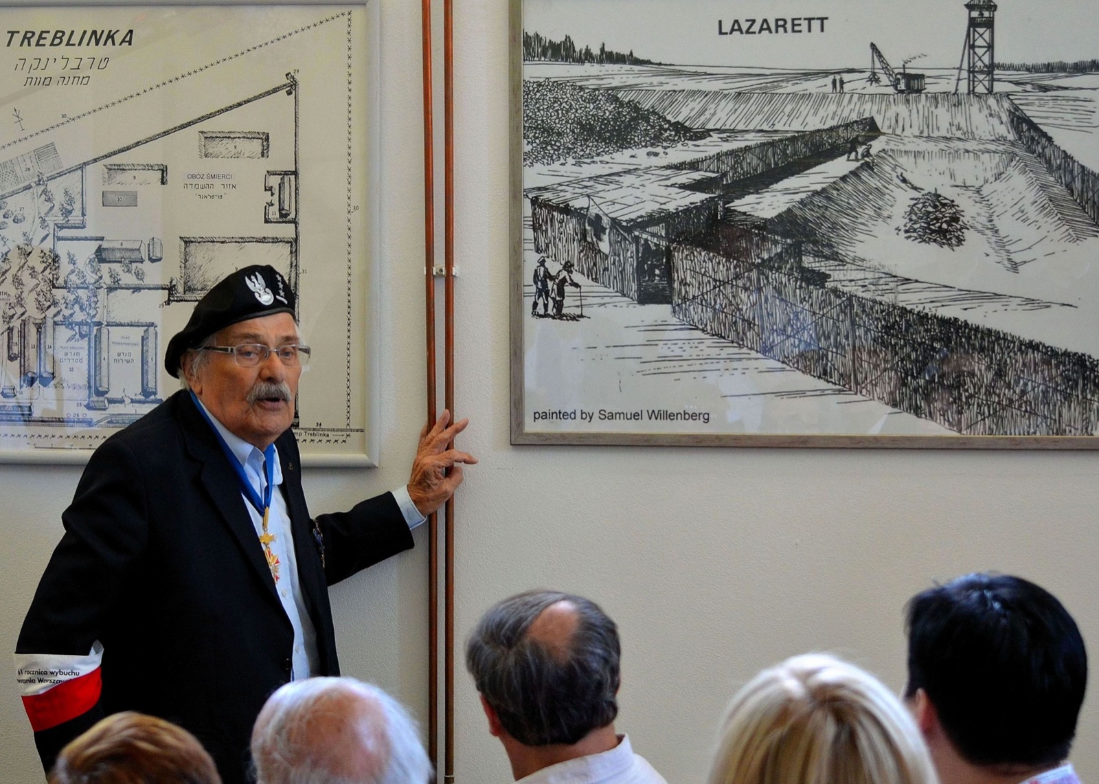 An elderly man in a light shirt stands beside display boards at an outdoor memorial site, gesturing toward them as people look on.