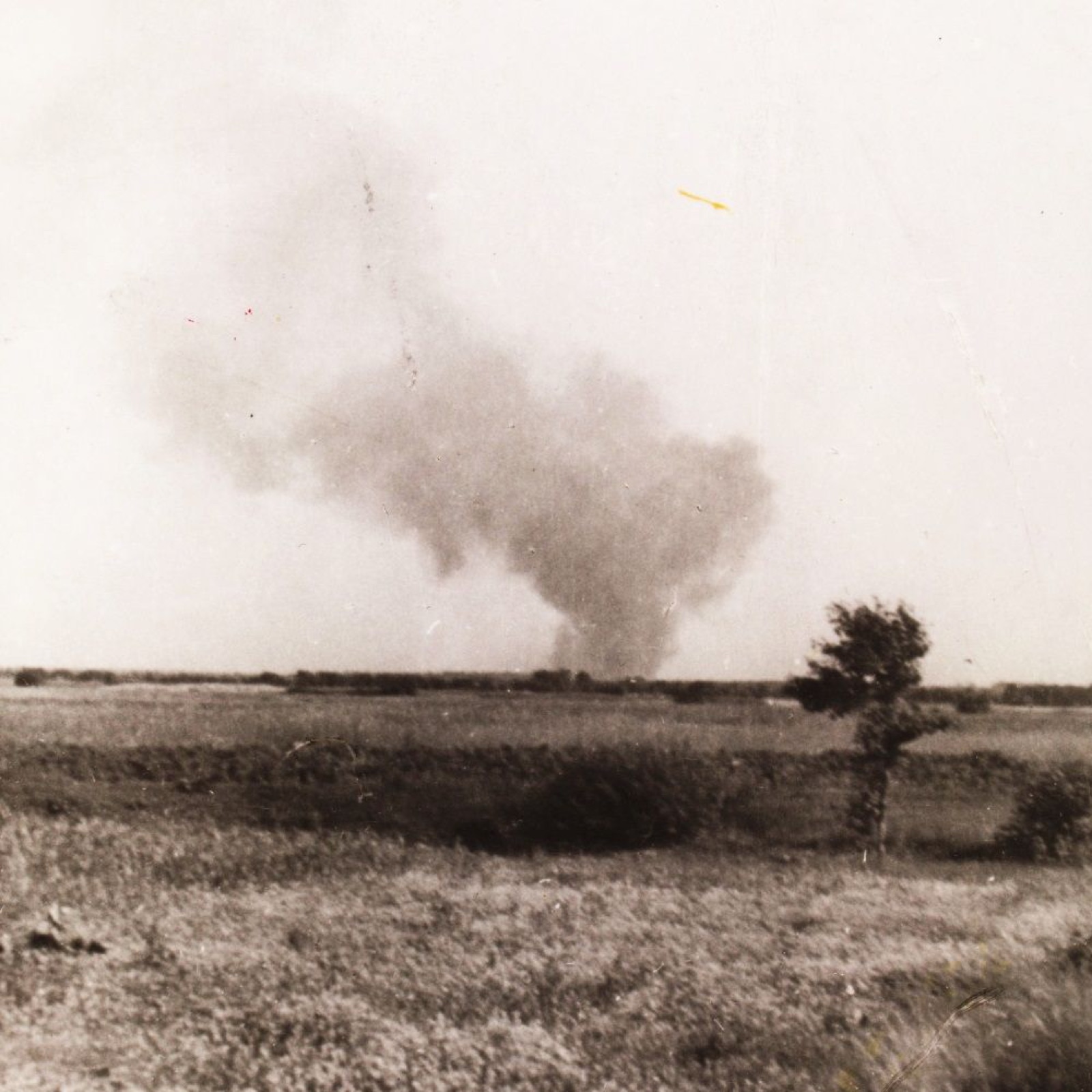 A distant view across flat country toward a low treeline, with a dark column of smoke rising above it into a pale sky.