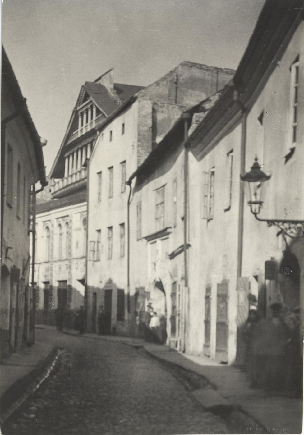 A narrow cobbled street of old plastered houses in Vilna, with a street lamp on the right, a few people standing in doorways, and a tall building with tiers of windows at the end of the street.