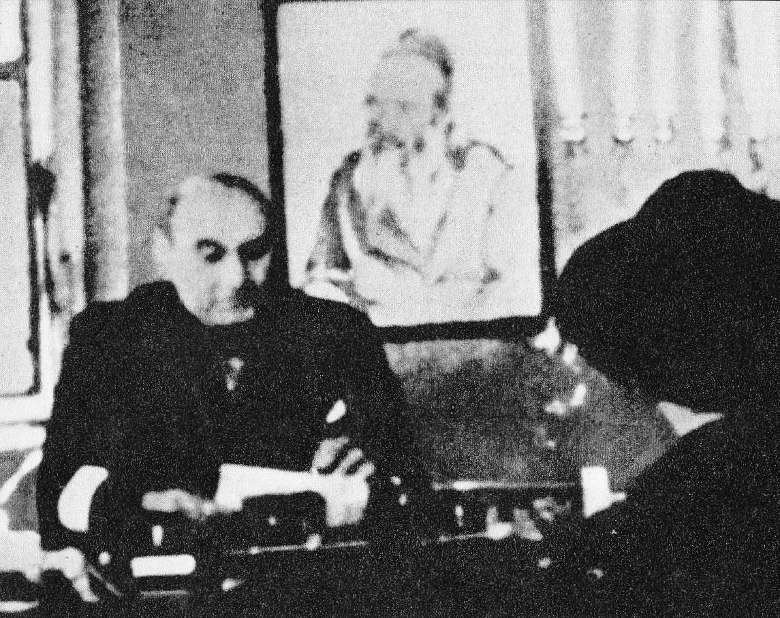 A man in a suit and tie seated behind a desk in an office, papers in front of him, facing the camera.