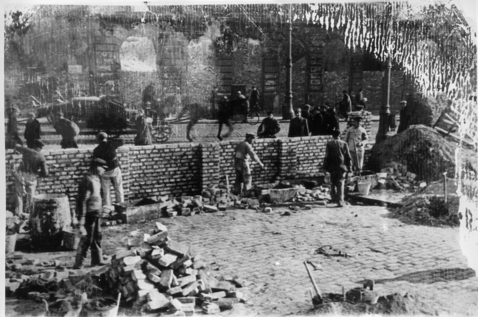 Bricklayers on a low scaffold raising an unfinished brick wall across a city street. Traffic and pedestrians move along the open roadway behind them, and a bomb-damaged building stands at the corner.