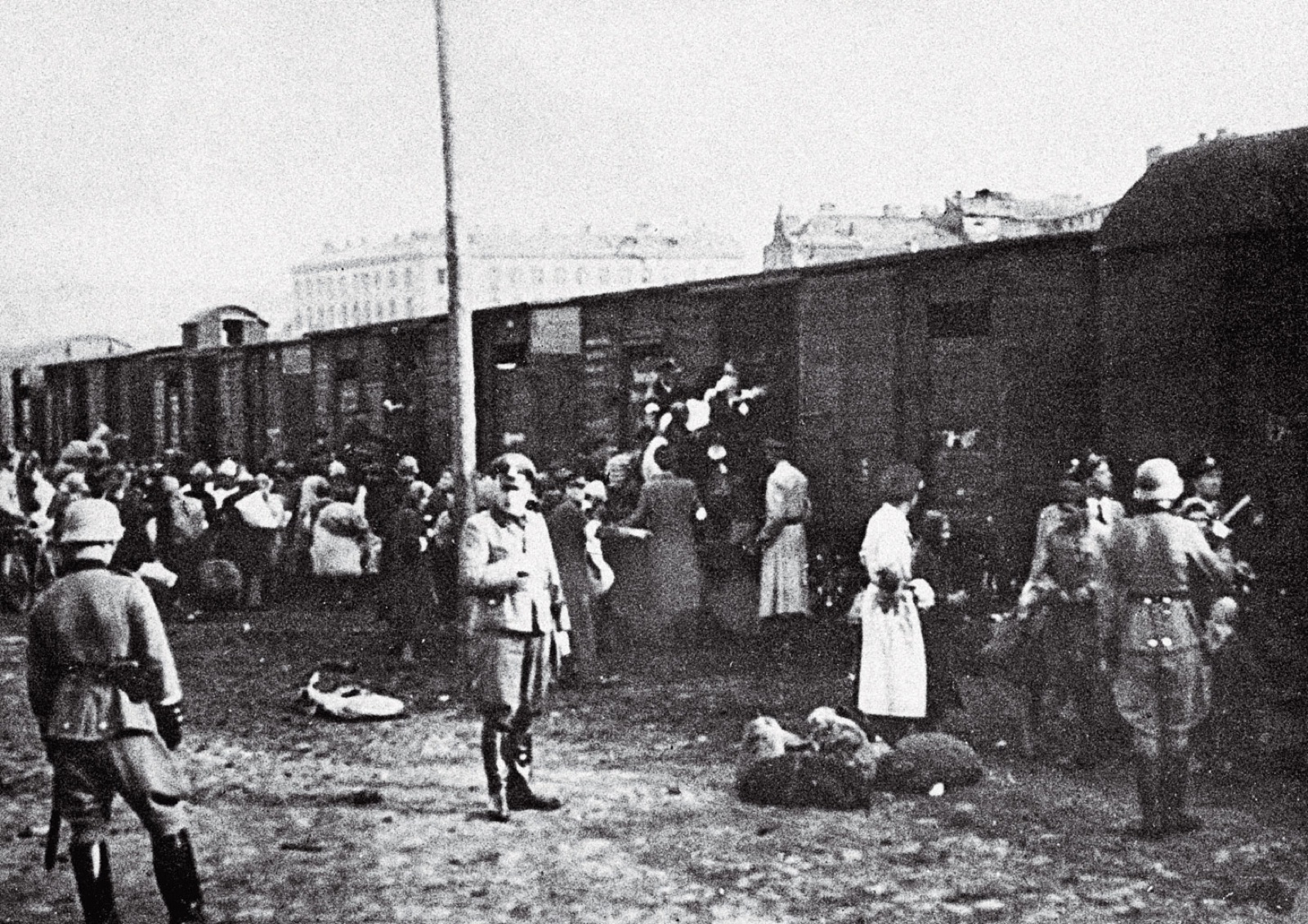People carrying bundles being pressed toward the open doors of wooden freight wagons at a rail siding. Children are in the crowd.