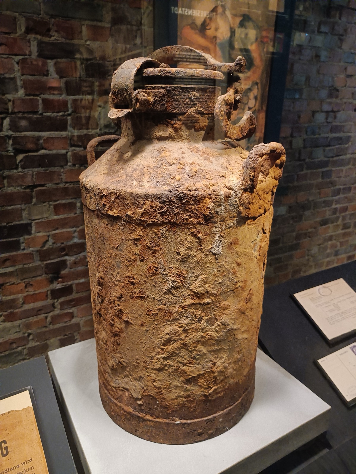 A dented metal milk churn with a lid and two side handles, standing alone under museum lighting.