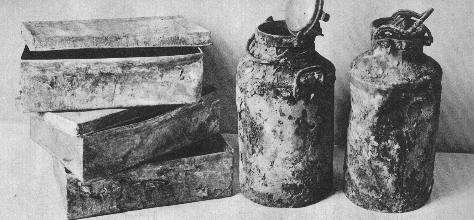 Three dented metal boxes and two tall milk cans standing side by side on a plain surface.