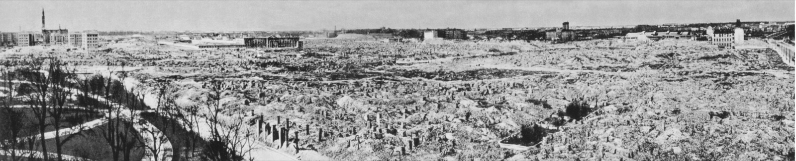 A wide panorama of open ground where a city district stood, low mounds of brick rubble running back to a line of surviving buildings on the horizon.