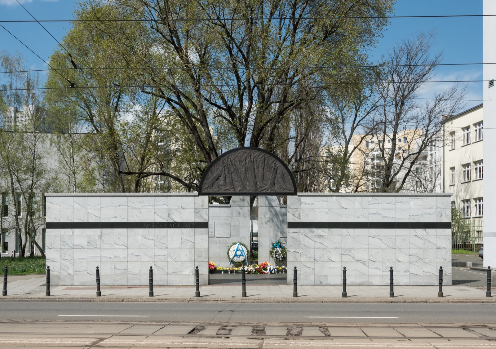 A low monument of pale stone standing open at the center like a gateway, in a paved square, its inner faces covered in columns of engraved names.