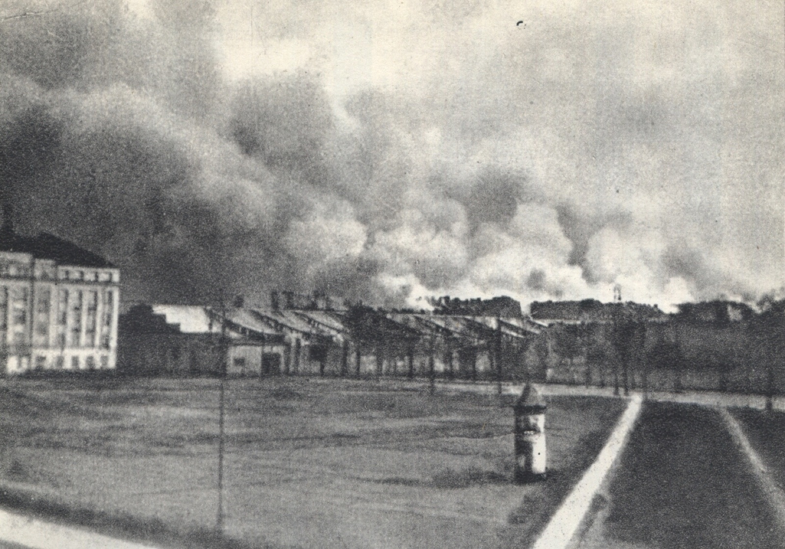 A city skyline at night seen across low rooftops, with a wide band of smoke and firelight rising from a district in the middle distance. No people are visible.