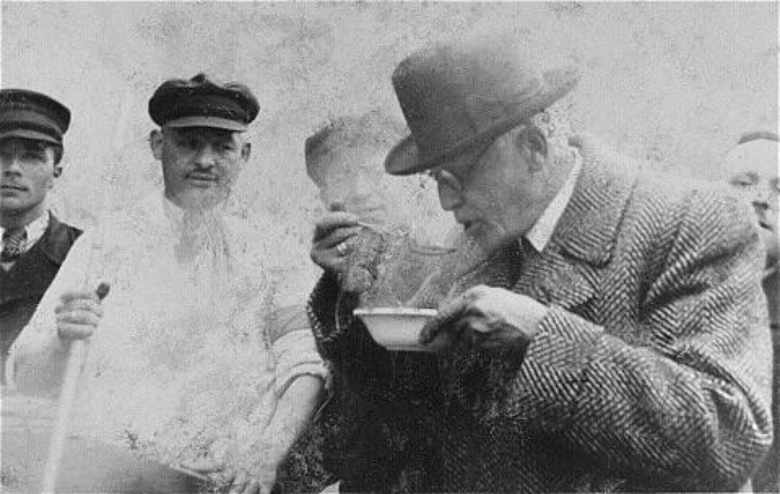 A middle-aged man in a coat leans over a large cooking vessel with a spoon raised to his mouth while several people in aprons watch him.