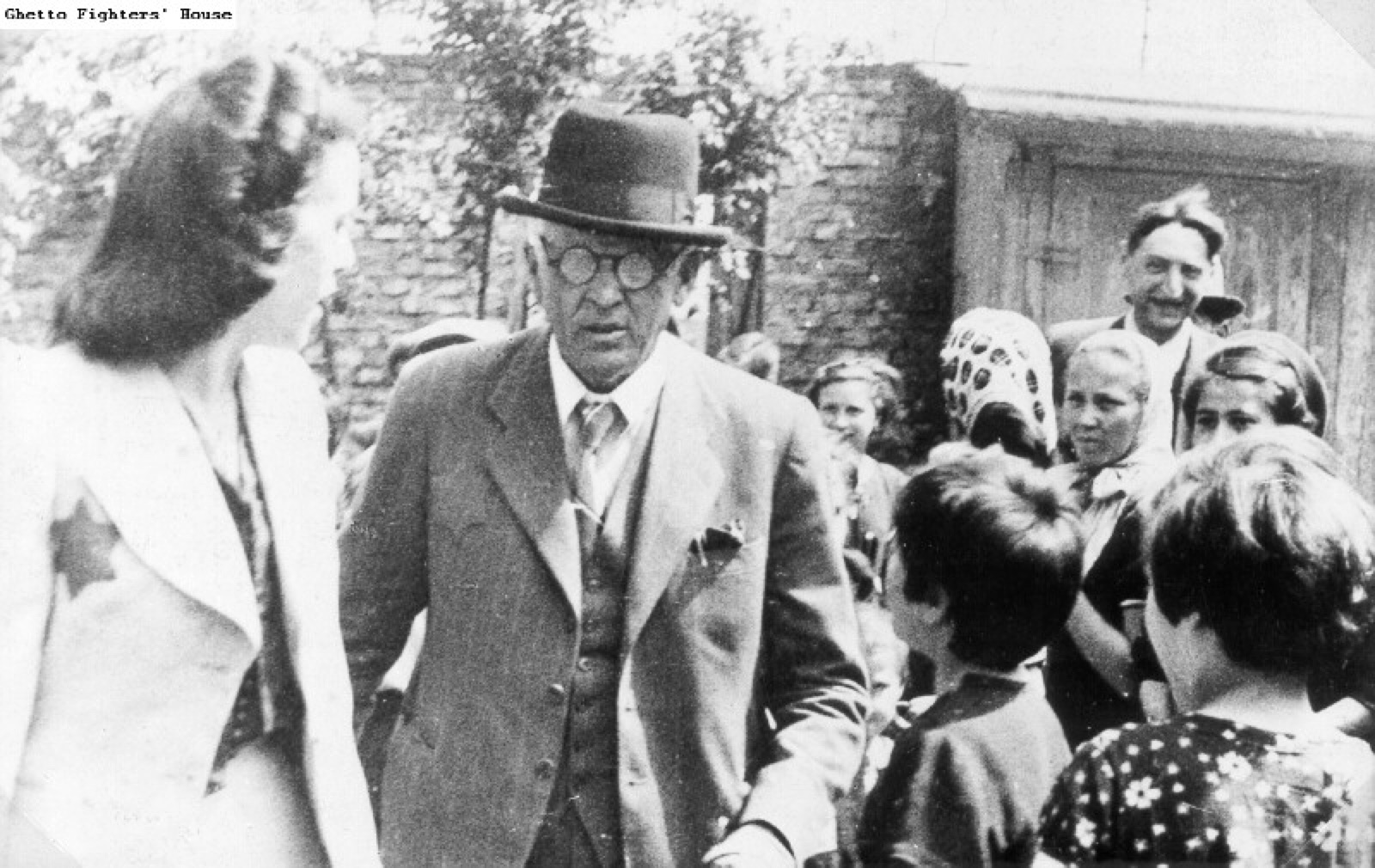 A man in a coat and cap stands outdoors among a group of children of about school age, some of them turned toward him and some toward the camera.
