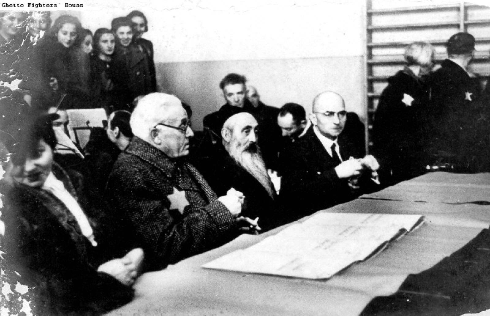 An indoor ceremony. Adults sit at a table at the front, with rows of young people standing and seated behind them.