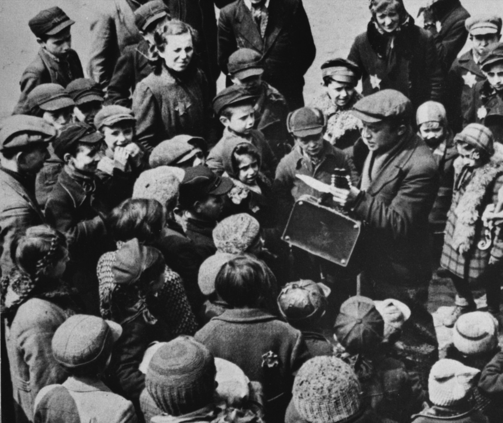 A man in a jacket and cap stands singing in the open air, his mouth open and one hand lifted, with a few people behind him.