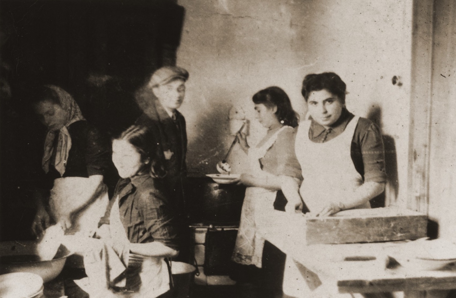 Workers in aprons standing over large cooking vessels in a communal kitchen, serving bowls stacked on the counter beside them.