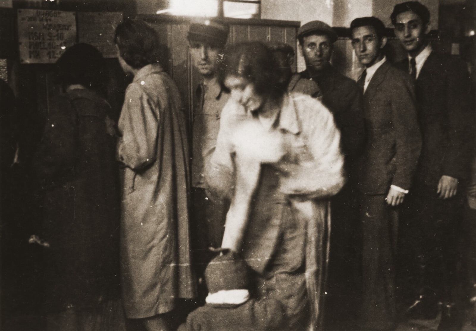 A line of people inside an office in the Lodz ghetto: men in caps and coats along the wall, two young men looking at the camera, a woman in the foreground bending over a bundle.