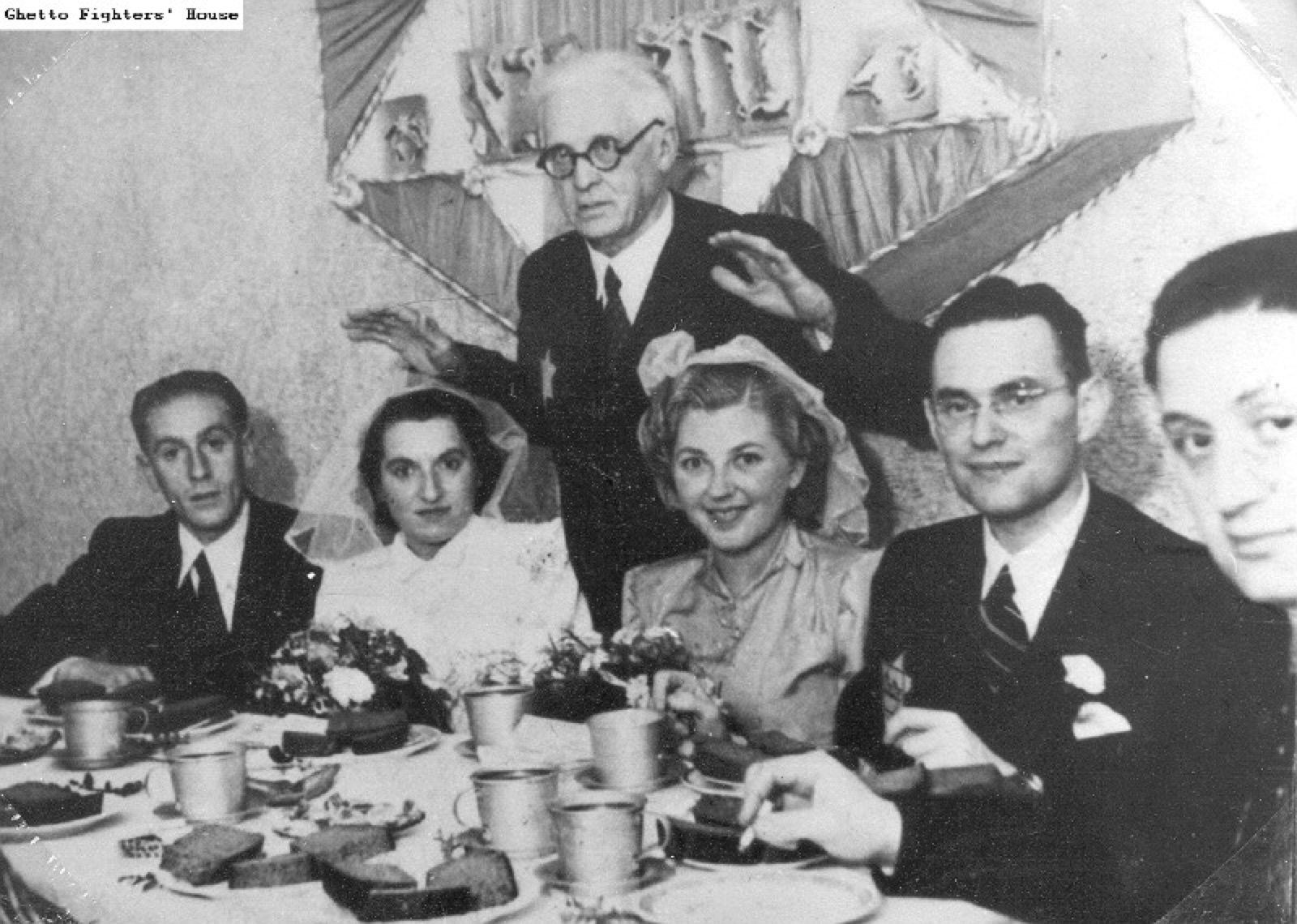 Two couples standing side by side while an older man in a heavy coat holds his hands out over them.