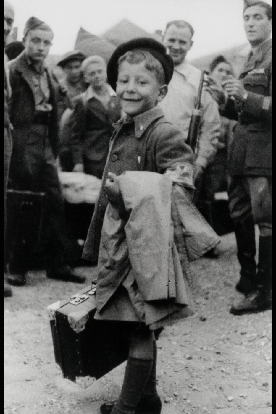 An eight-year-old boy carrying a suitcase walking away from Buchenwald, with an older boy behind him.