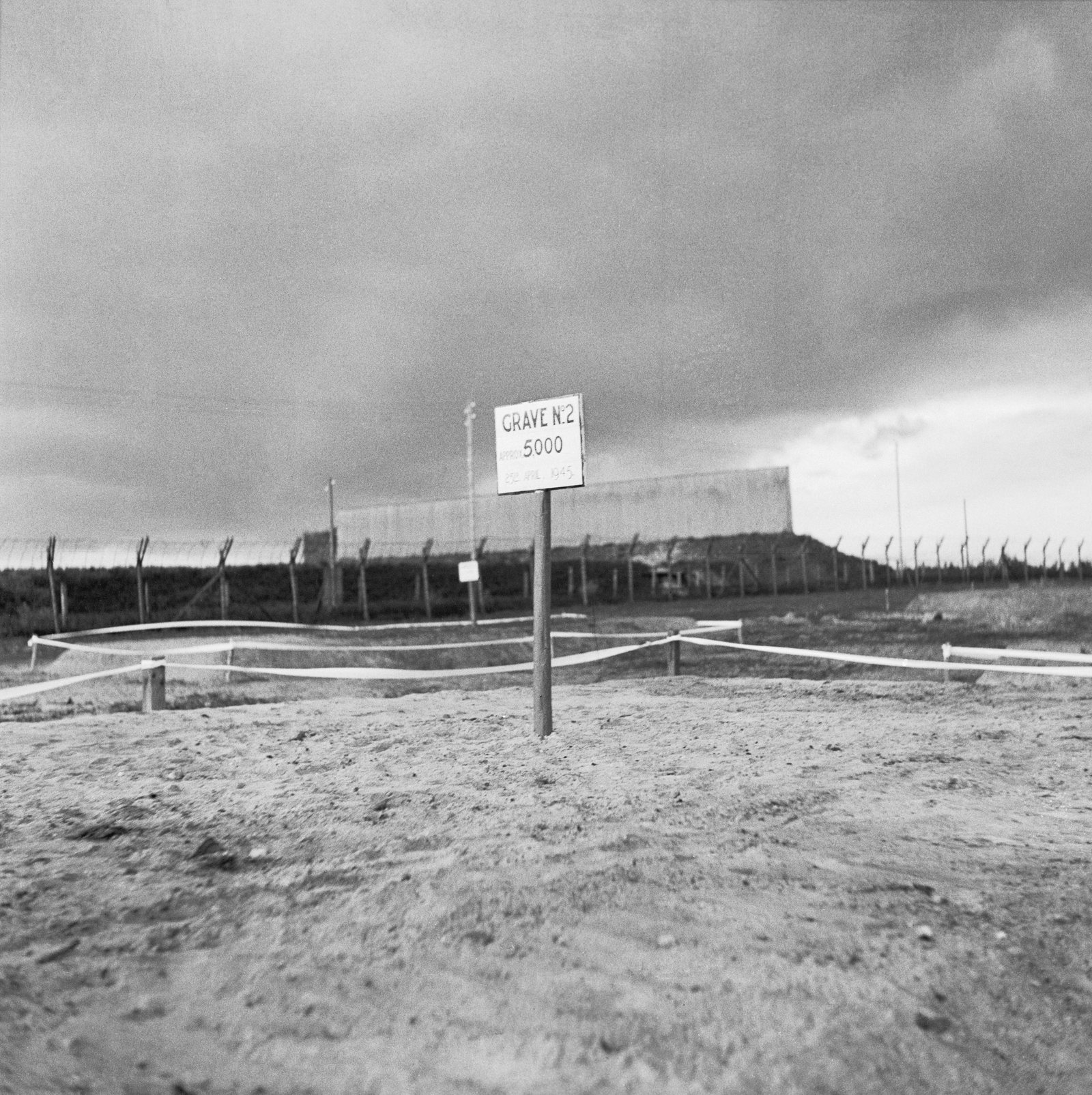 A wooden sign on a post in bare, flattened sand, reading Grave No 2, approx. 5,000, with a rope barrier around the grave and a barbed-wire fence and huts under a dark sky behind it.