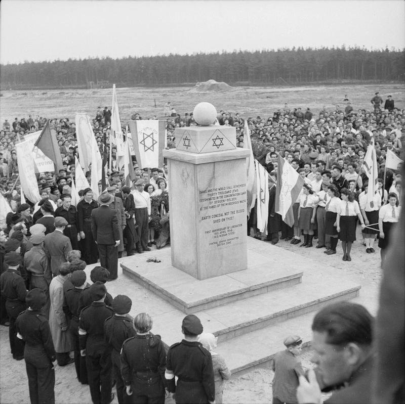 A crowd of survivors with banners and Star of David flags around a tall stone memorial at Bergen-Belsen, with a line of uniformed youth in the foreground and a pine forest beyond the open ground.