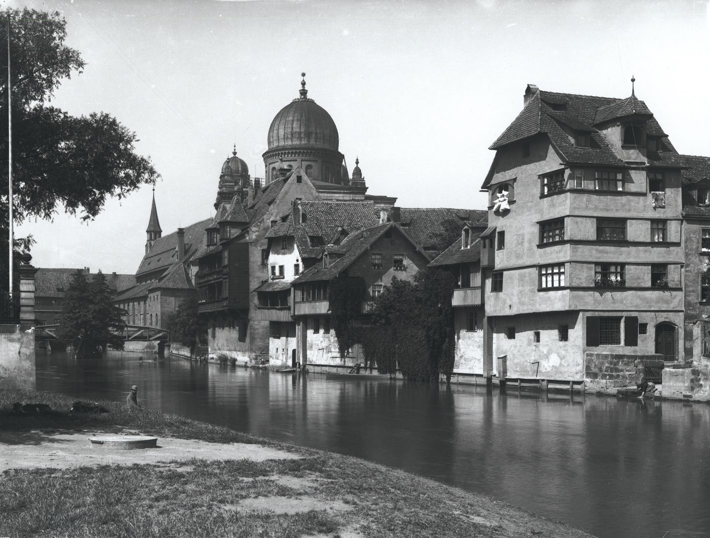 The domed synagogue of Nuremberg rising behind old half-timbered houses on the bank of the river Pegnitz, with a tree and a riverside path in the foreground, about 1900.