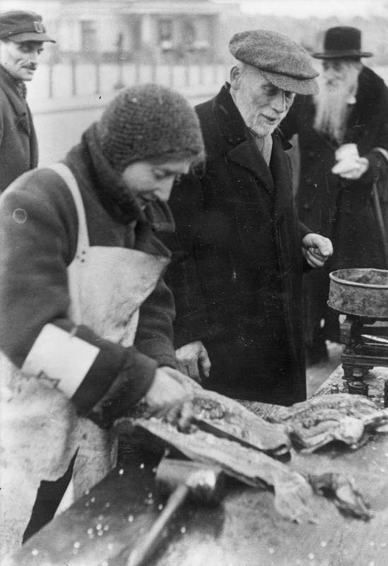 Black and white photograph of a woman in an apron and armband selling fish at an outdoor stall, watched by two elderly men
