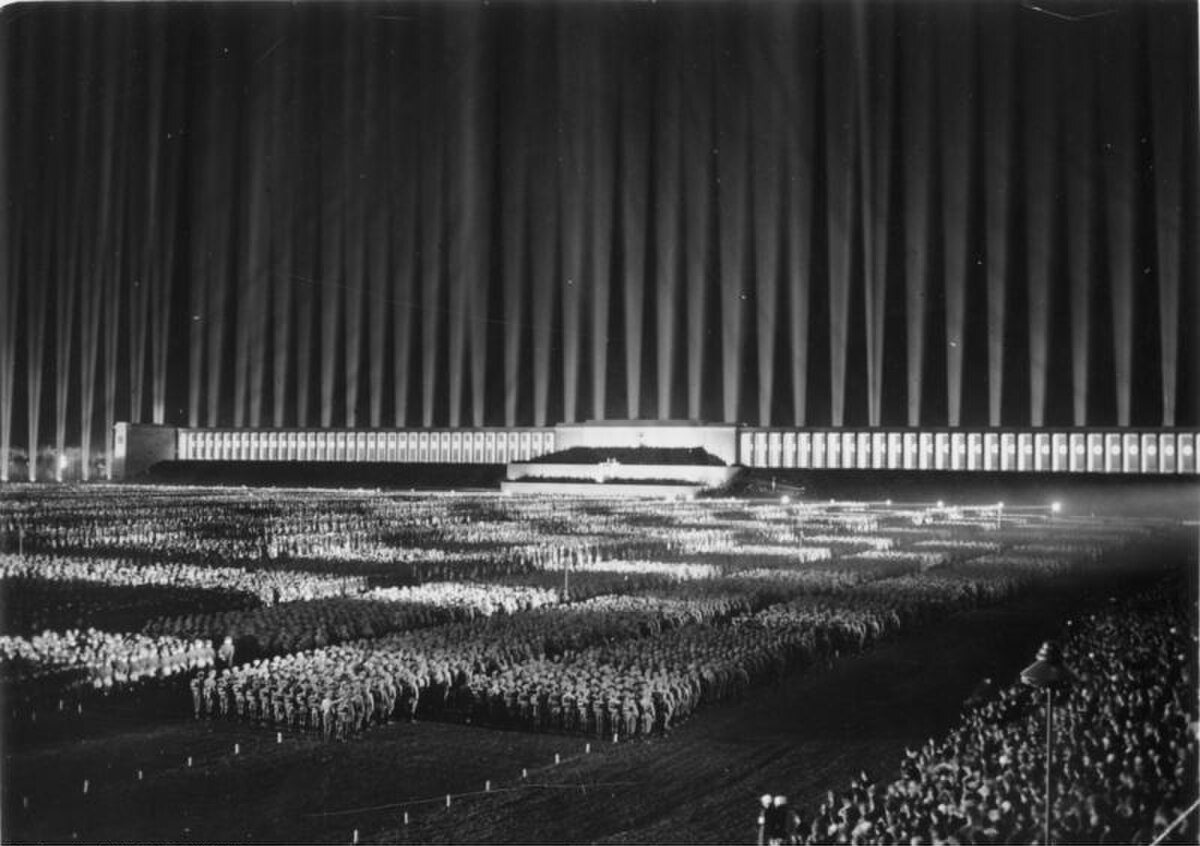 Night photograph of vertical searchlight beams rising above a rally ground, forming columns of light
