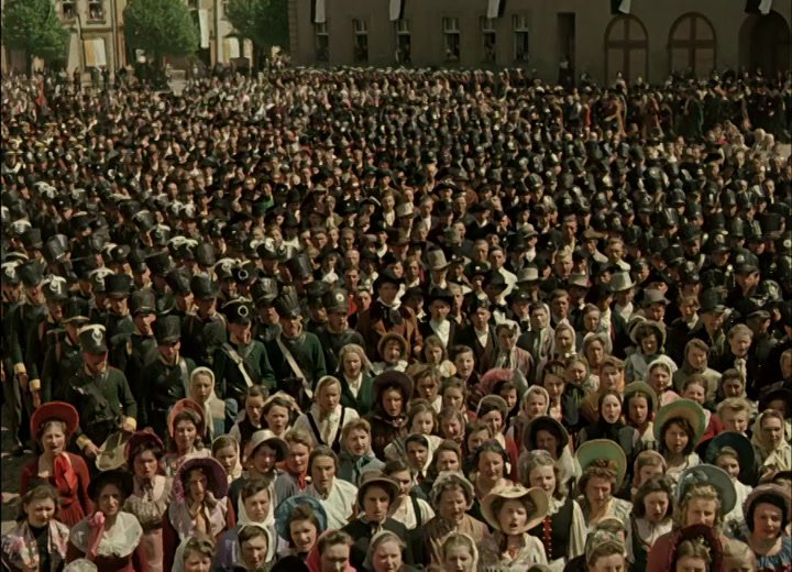 Colour film still of a period crowd scene with soldiers and townspeople
