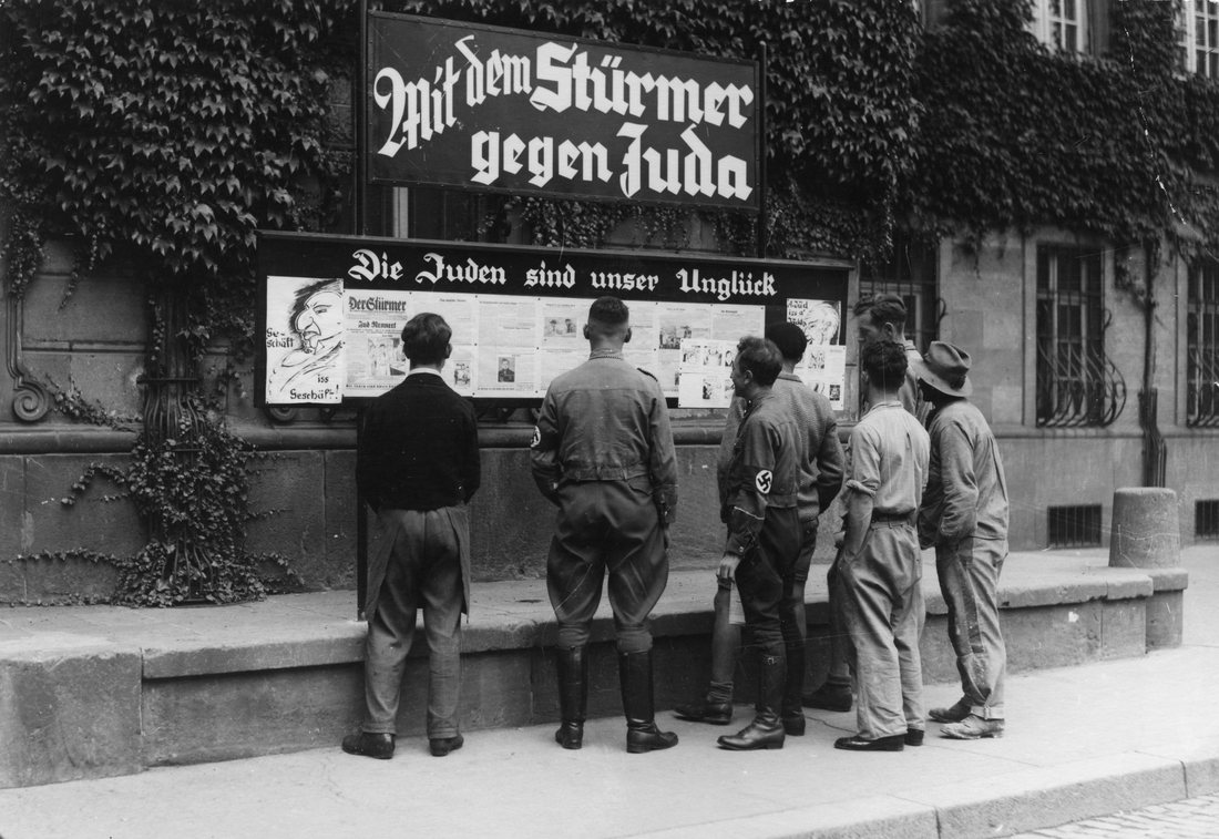 People standing at a street display case reading a mounted newspaper under a German-language banner