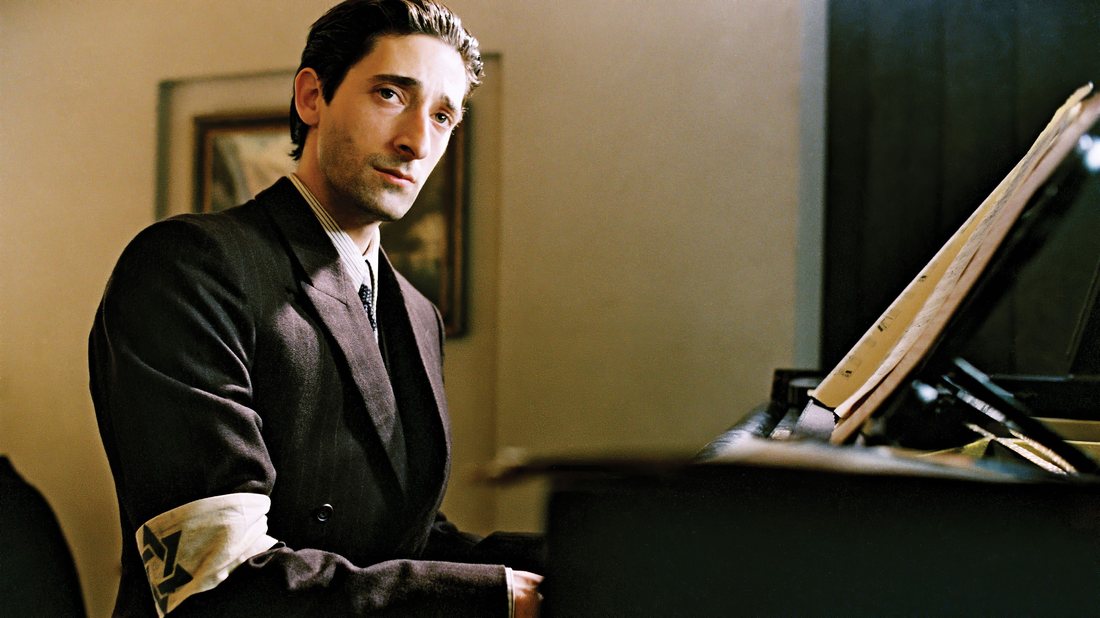 A gaunt man in a dark suit seated at a grand piano in a ruined room