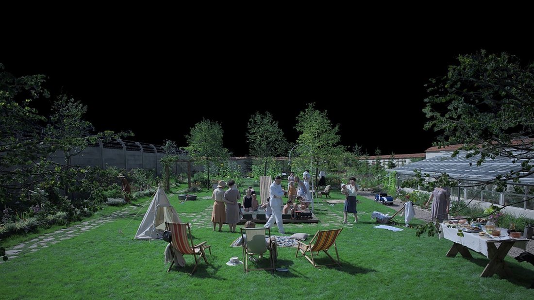 A manicured garden with deck chairs and people relaxing, directly against the high wall of a concentration camp