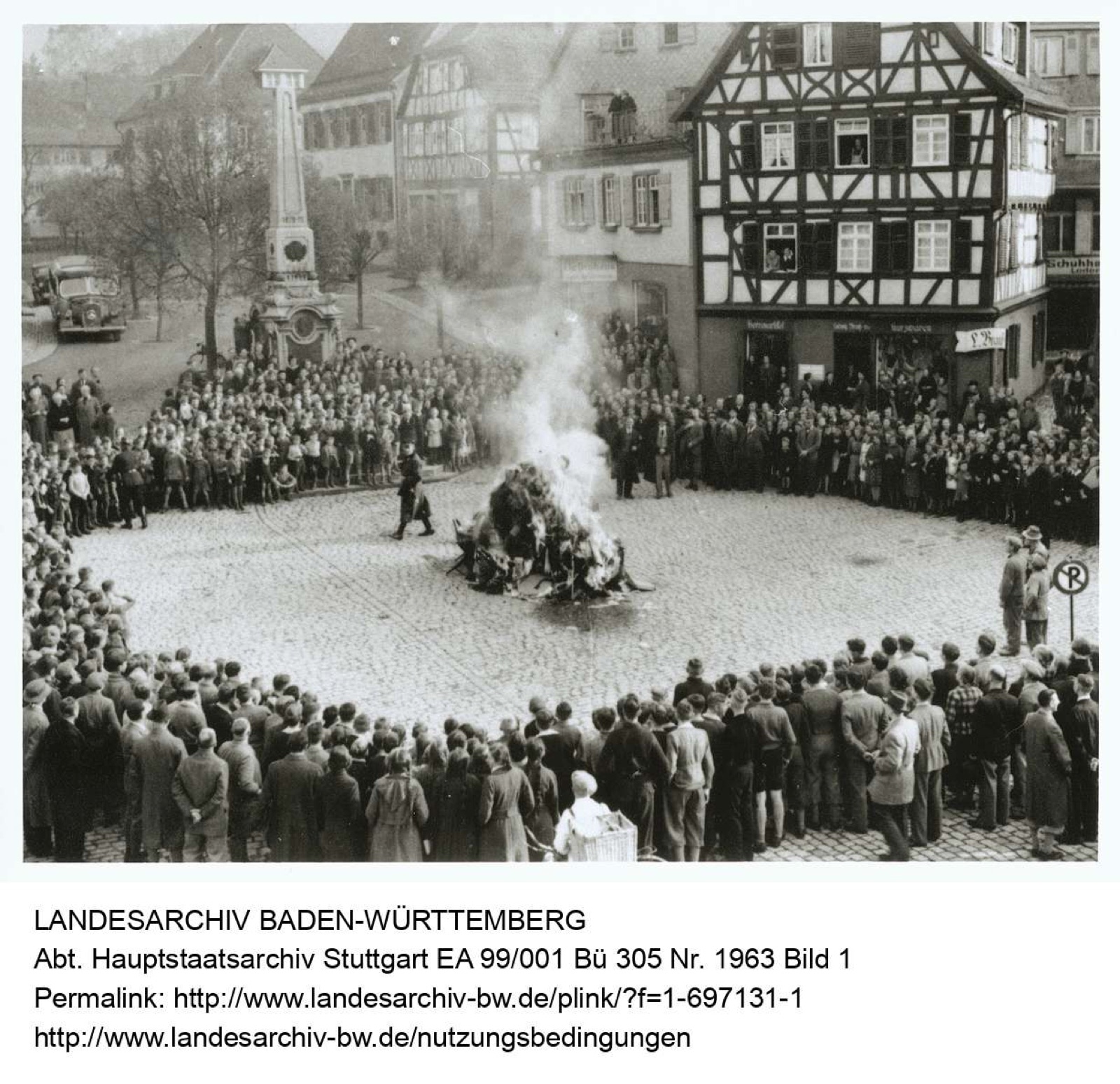 A dense crowd of men, women and children filling a small-town street, most in winter coats and hats, all facing the same direction; buildings with shuttered upper windows stand behind them.
