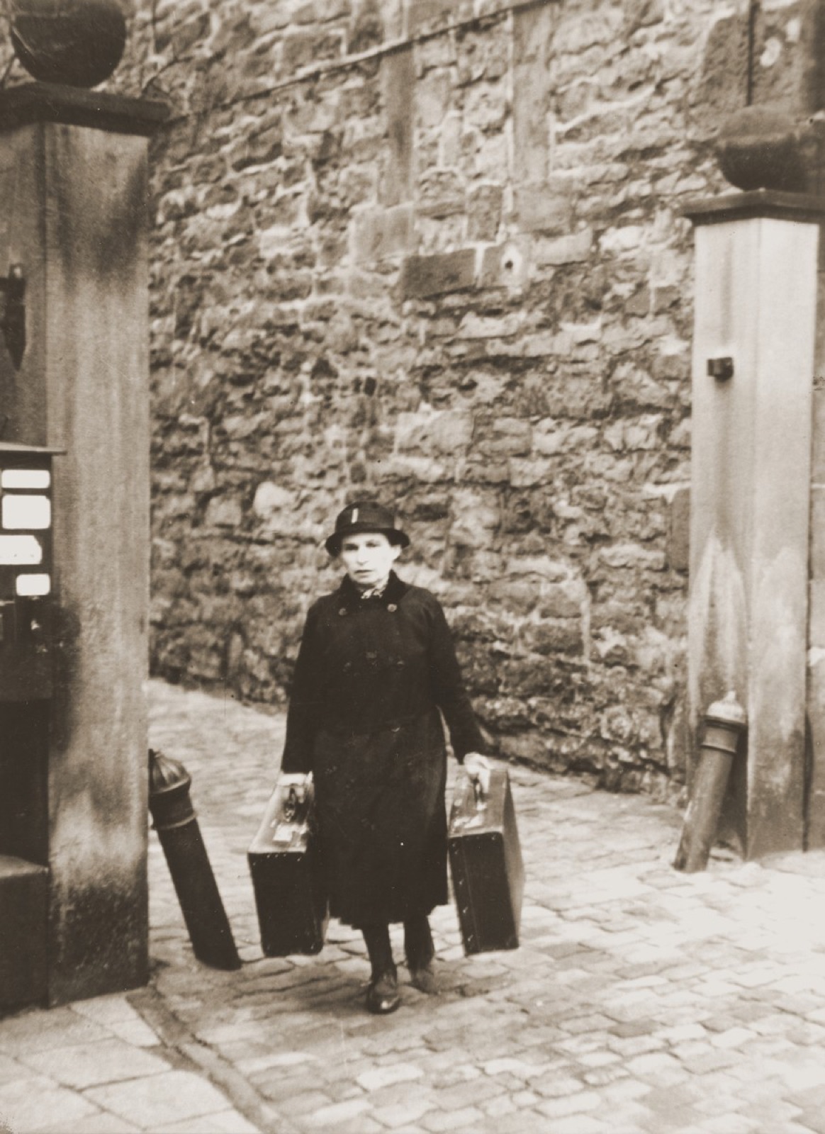 A woman in a coat standing in the open doorway of a house, a suitcase held in each hand, about to step down into the street.
