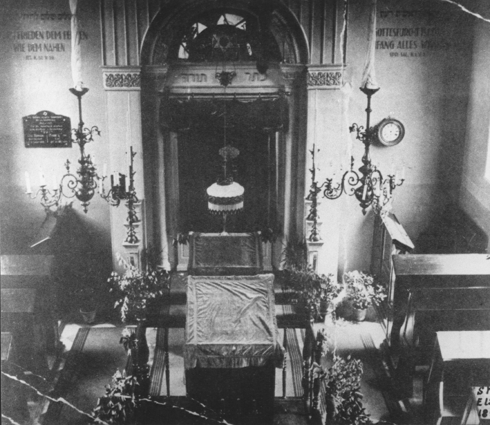 The interior of a small synagogue looking toward the ark, rows of wooden benches, a raised bimah at the center and a lamp hanging above it.