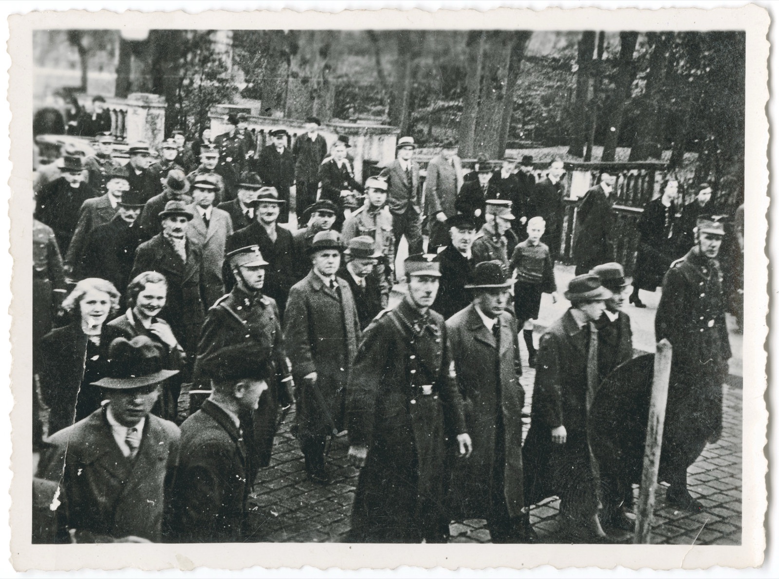 A double file of men in overcoats and hats walking down a paved street between uniformed escorts, with townspeople standing on the sidewalk on both sides watching them pass.