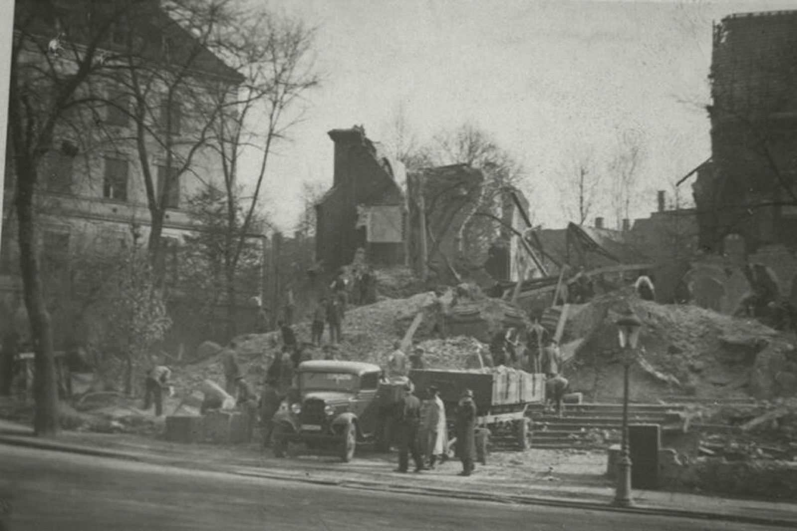 Several men in work clothes and caps standing on a heap of broken masonry and roof timbers, the shell of a large building behind them.