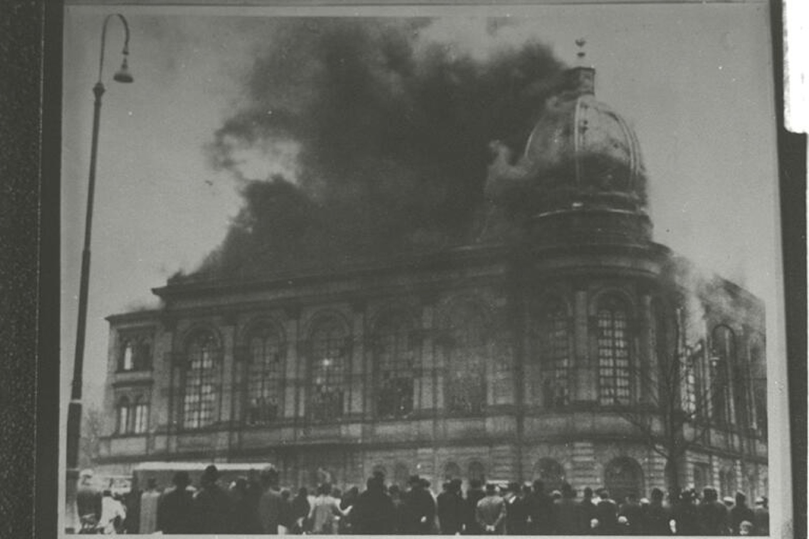 A large synagogue on fire, smoke and flame coming through the roof and the upper windows, the street in front of it visible below.
