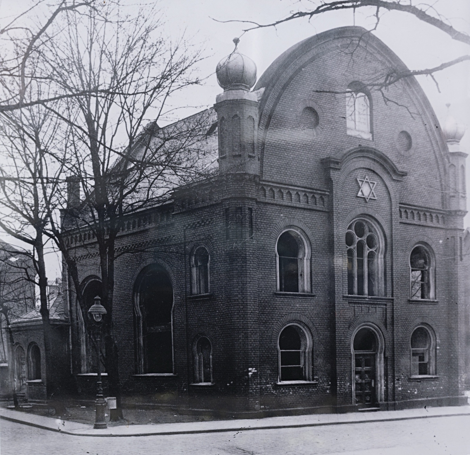 The burned out shell of a synagogue in daylight, the walls standing and the roof gone, seen from the street.