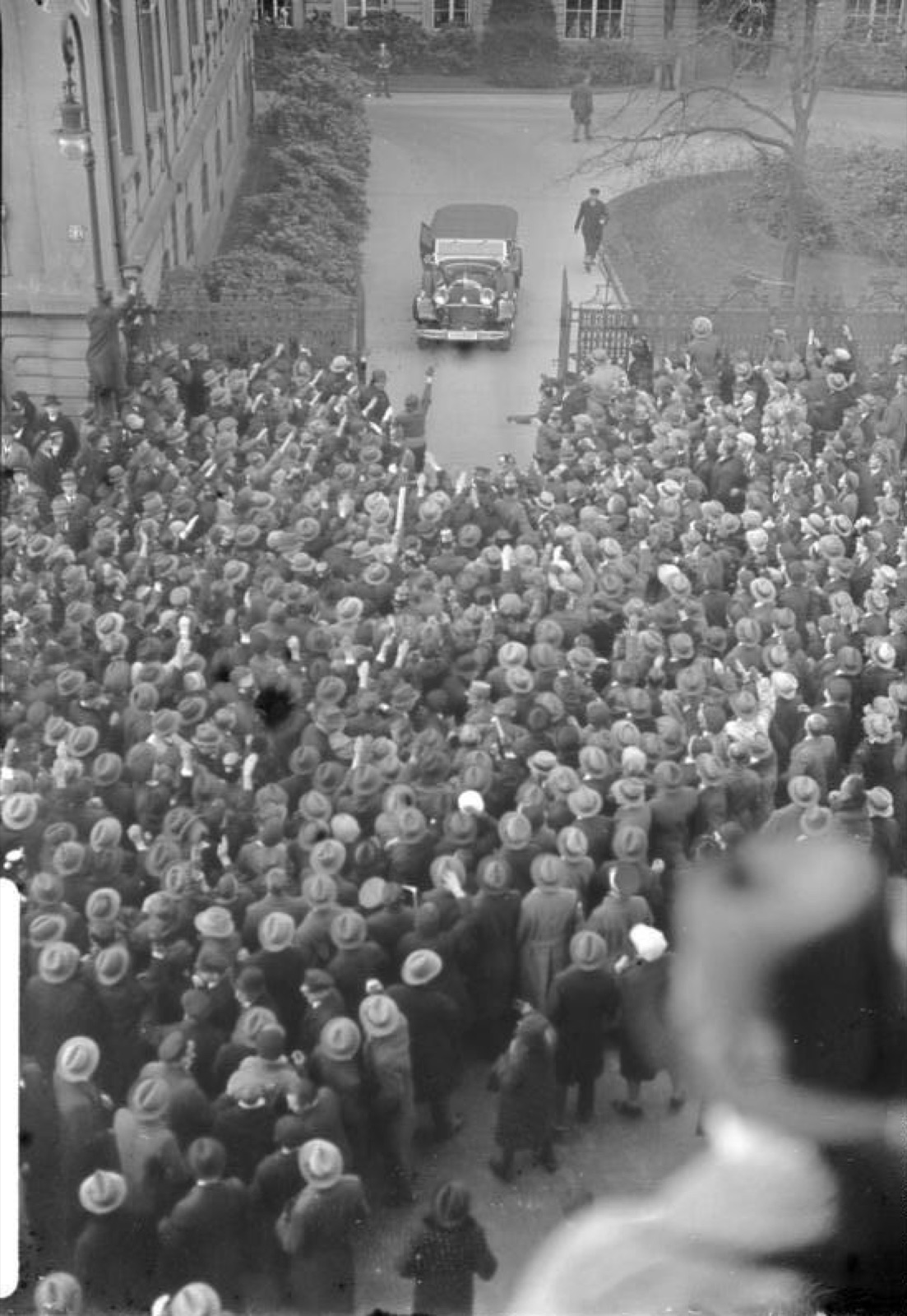 An open car pulling away from the Reich Chancellery on the Wilhelmstrasse, with a crowd pressing along the street.