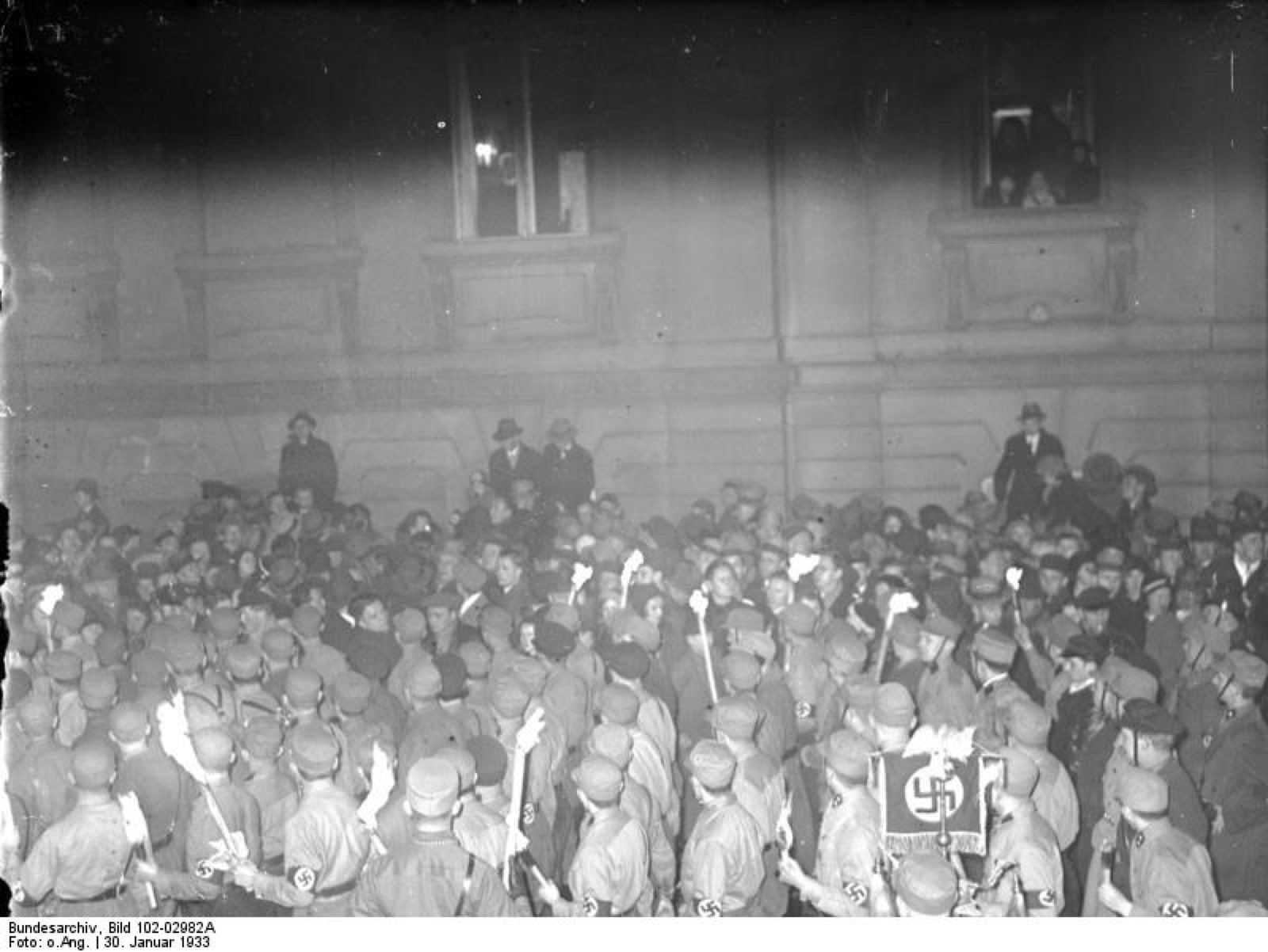 A torchlit column moving along a Berlin street at night, seen from the lit windows of the buildings above.