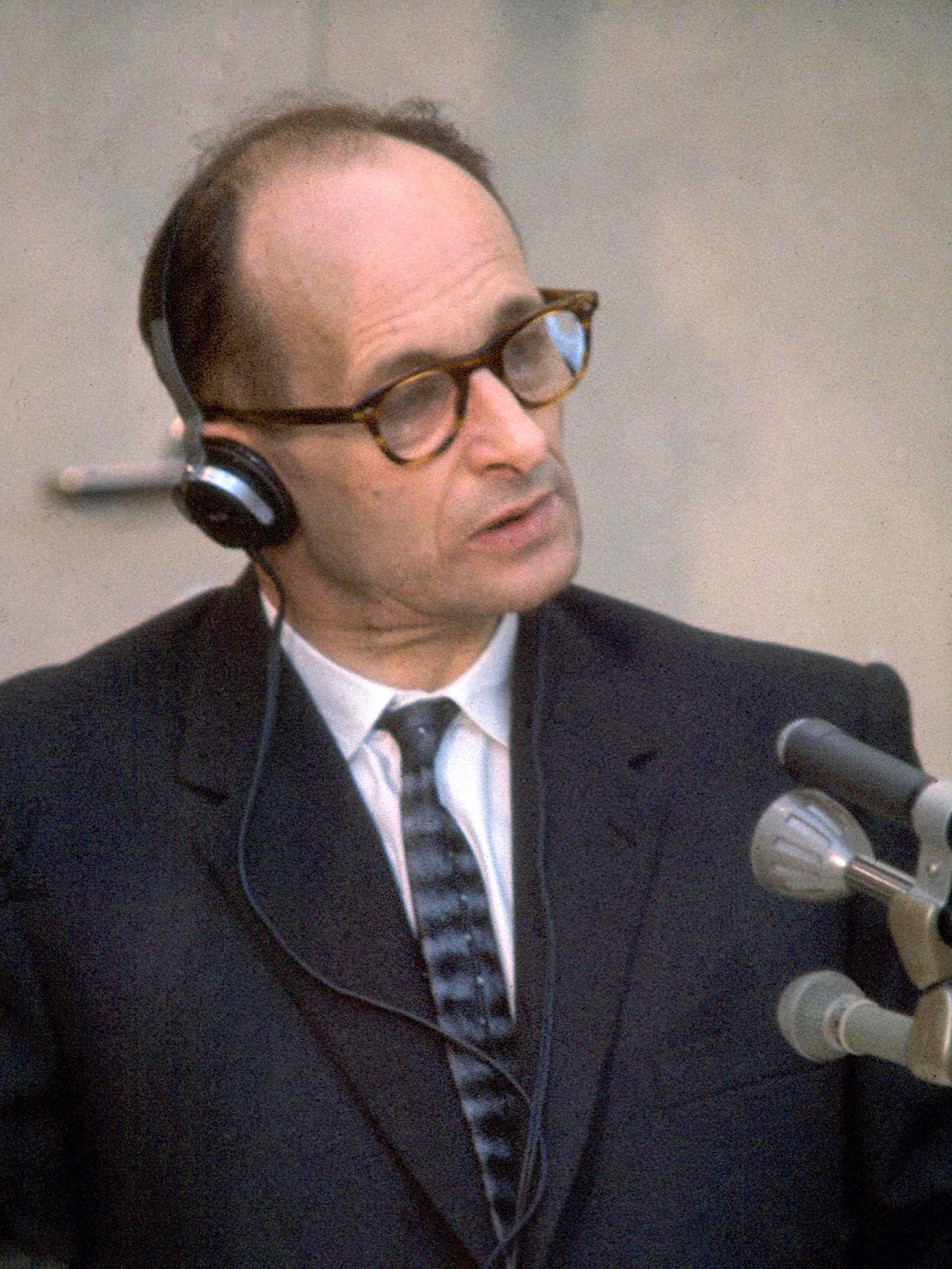 A balding man in a dark suit sits alone inside a booth of clear panels, headphones on, hands resting on a small table, with a uniformed guard visible behind the glass.
