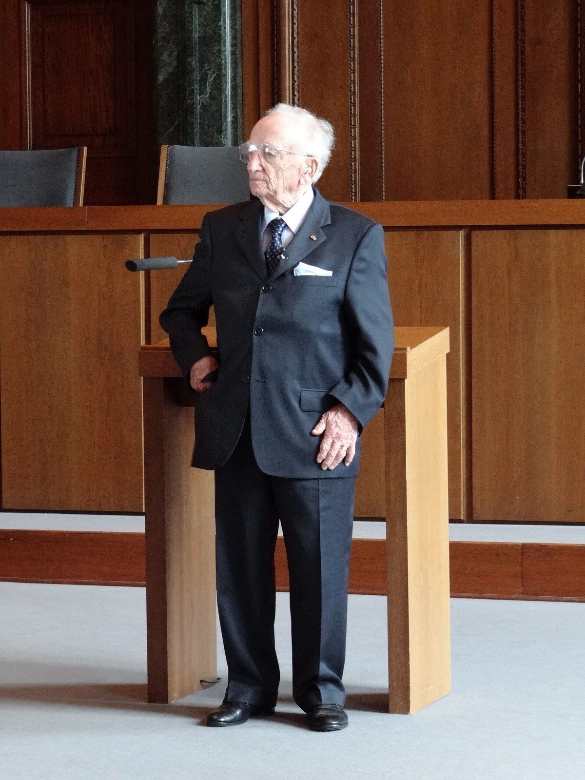 An elderly man in a dark suit stands alone in an empty wood-paneled courtroom, looking toward the camera.